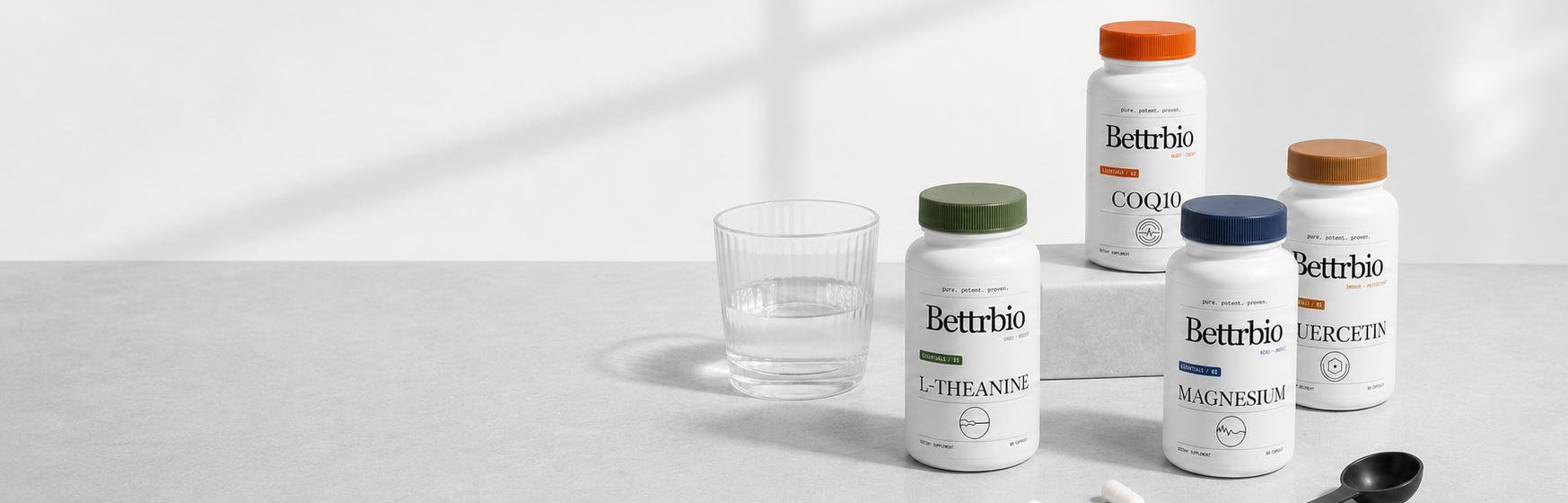 Bettrbio Essentials family—L-Theanine, Magnesium, CoQ10, and Quercetin—fully framed on a panoramic cool studio worktop with water, capsules, and a measuring spoon